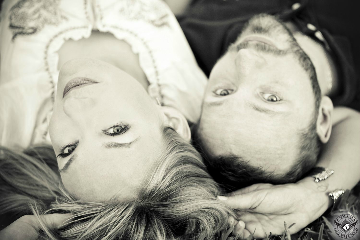 Smooth black and white of a blond woman with bright eyes in a ruffled white button up blouse lies on her back next to a dark haired bearded guy wearing a dark casual collared shirt in the grass at the Texas Capital in this romantic engagement portrait in Austin. 